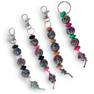 May include: Four keychains with silver-tone clasps and key rings. Each keychain features a strand of colorful beads, including leopard print spheres, pink ovals, and other shapes. The keychains are finished with decorative charms.