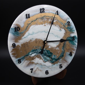 Può includere: Orologio da parete rotondo con quadrante bianco, con motivi astratti dorati, turchese e bianchi. I numeri neri segnano le ore e le lancette nere indicano l'ora. L'orologio è esposto su un supporto di legno.