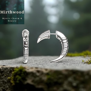 May include: Silver-toned earrings with a curved, claw-like design. One earring is shown upright, while the other is curved. The earrings have detailed patterns and the text "Mirthwood Mystic Charm & Beauty" is in the upper left corner.