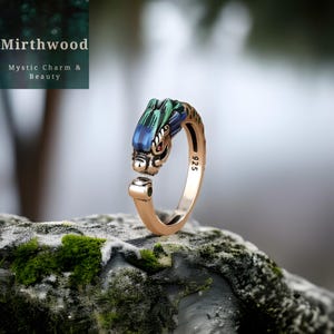 May include: A gold-colored ring with a dragon head design. The dragon's head features blue and green accents, with a red gemstone eye. The ring is set on a mossy rock, with the text "Mirthwood Mystic Charm & Beauty" in the background.