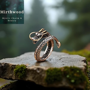 Könnte beinhalten: Ein bronzefarbener Drachenring mit aufwendigen Details. Kopf und Schwanz des Drachens umschließen den Finger, mit einem strukturierten Schuppenmuster. Der Ring liegt auf einer Stein Oberfläche, mit dem Text "Mirthwood Mystic Charm & Beauty".