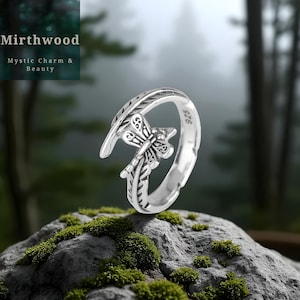 May include: A silver ring with a dragonfly design. The ring features a detailed dragonfly and feather motif. The ring is set against a natural background with the text "Mirthwood Mystic Charm & Beauty".