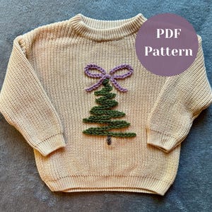 Könnte beinhalten: Beigefarbener Strickpullover mit einem grünen Weihnachtsbaum und einer lila Schleifenapplikation. Der Pullover hat einen runden Halsausschnitt und lange Ärmel. Der Text "PDF Pattern" befindet sich in einem lila Kreis.