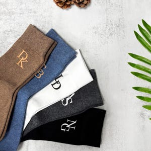 Può includere: Un assortimento di cinque paia di calzini in vari colori: marrone, blu, bianco, grigio e nero. Ogni calzino presenta un'iniziale ricamata diversa. I calzini sono disposti per mostrare i loro colori e disegni.