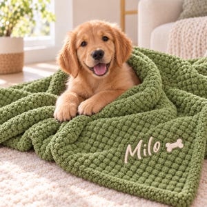 Puede incluir: Un cachorro de golden retriever descansando sobre una manta de color verde oliva con textura. La manta presenta un diseño bordado personalizado en hilo blanco que dice Milo junto a un pequeño icono en forma de hueso.