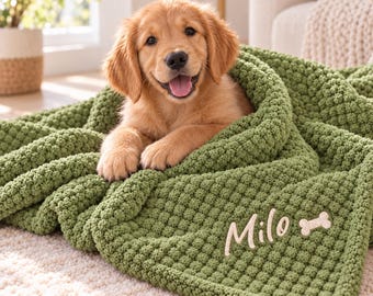 May include: A golden retriever puppy resting on a textured olive green blanket. The blanket features a personalized embroidered design in white thread that reads Milo next to a small bone-shaped icon, adding detail to the fabric.