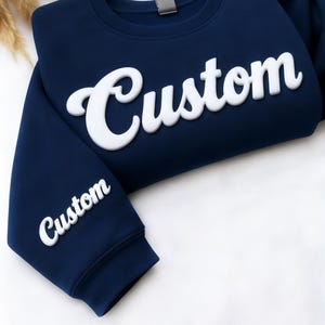 May include: Navy blue sweatshirt with the word "Custom" in large, white, puffy letters across the chest. The word "Custom" is also printed on the left sleeve. The sweatshirt is made of a soft material.