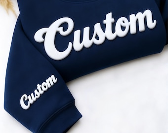 May include: Navy blue sweatshirt with the word "Custom" in large, white, puffy letters across the chest. The word "Custom" is also printed on the left sleeve. The sweatshirt is made of a soft material.