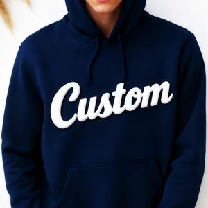 May include: Navy blue hoodie with the word "Custom" in white, stylized lettering across the chest. The hoodie features a hood with drawstrings and a front pocket. The garment is made of a soft material.