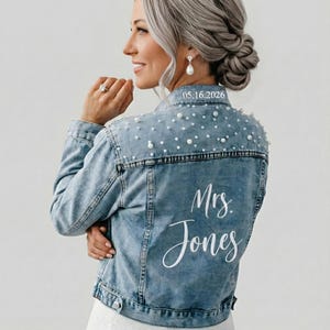 May include: Pale blue denim jacket adorned with pearls. The back of the jacket displays the text "Mrs. Jones" in white script, with the date "05.16.2026" above. A stylish bridal or wedding jacket.