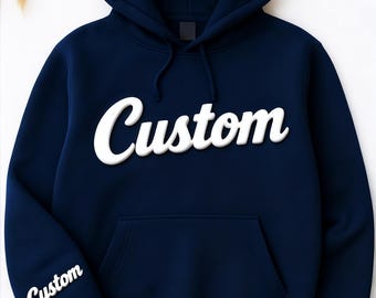 May include: Navy blue hoodie with the word "Custom" in white, bold, cursive lettering across the chest. The same word is also printed on the left sleeve. The hoodie has a hood and a front pocket.