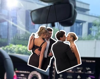 May include: Car air fresheners featuring images of couples in formal attire. One shows a couple kissing, the other a couple embracing, with a champagne glass. The background includes a car's interior and a building.