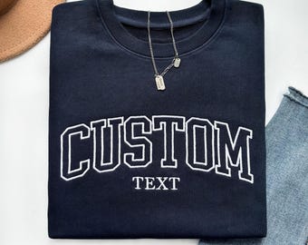 Może przedstawiać: Granatowa bluza z napisem "CUSTOM" w białych, łukowatych literach i "TEXT" poniżej. Srebrny naszyjnik z dwiema małymi zawieszkami spoczywa na bluzie. Widoczna jest para niebieskich dżinsów. Bluza wykonana jest z miękkiego materiału.