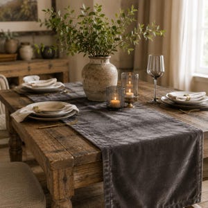 May include: A dark gray velvet table runner laid across a rustic wooden dining table. The table is set with ceramic plates, linen napkins with gold-toned rings, a tall glass vase with green branches, two glass candle holders, and a wine glass.