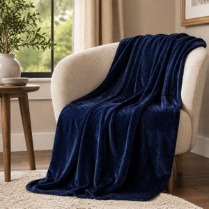 Pode incluir: Uma manta de pelúcia azul marinho sobre uma poltrona creme. A textura macia da manta é visível, com dobras e sombras que adicionam profundidade. A cadeira está sobre um tapete de tons neutros, com uma pequena mesa lateral de madeira e um vaso com vegetação ao fundo.