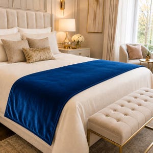 May include: A rectangular royal blue velvet bed runner draped across a cream-colored bedspread. The runner features a smooth, solid-colored fabric surface with clean, straight edges, providing a bold contrast against the neutral bedding.