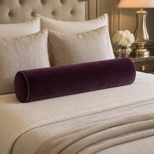 May include: A long, cylindrical bolster pillow upholstered in deep purple velvet fabric. The pillow features a smooth, solid-coloured surface with piped edges at both circular ends, positioned horizontally across the centre of a bed with neutral bedding.