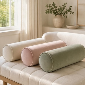 May include: Three cylindrical bolster pillows arranged in a row on a tufted bench. The pillows are made of a soft, solid-colored fabric in shades of cream, light pink, and sage green, each featuring a smooth, rounded shape and piped edges.