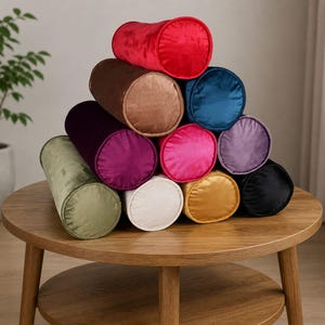 Op de afbeelding: Een verzameling cilindrische fluwelen kussens in verschillende kleuren, waaronder rood, bruin, blauwgroen, roze en zwart, gerangschikt in een piramidevorm op een houten tafel. De kussens hebben een zacht, pluche uiterlijk, wat duidt op comfort en stijl.