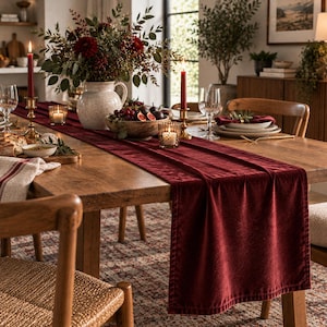 May include: A deep red velvet table runner draped over a wooden dining table. The runner features a soft, textured fabric surface with finished edges, extending across the length of the table beneath a centerpiece of flowers and candles.
