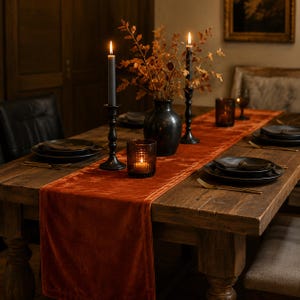 Puede incluir: Un camino de mesa de terciopelo color óxido centrado en una mesa de comedor de madera, con dos candelabros de metal negro con velas cónicas grises encendidas, un jarrón negro con follaje seco y un pequeño portavelas de cristal con una vela.