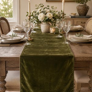 May include: A dining table set for a formal occasion. The table is covered with a dark green velvet runner, and set with white plates, crystal wine glasses, and gold-toned silverware. A floral arrangement and candles complete the elegant setting.