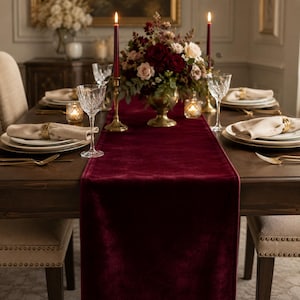 May include: A deep burgundy velvet table runner centered on a dark wooden dining table. The table is set with white plates, beige napkins held by gold rings, crystal glassware, gold cutlery, and two tall burgundy taper candles in gold holders.