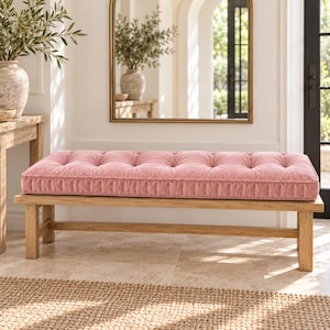 May include: A rectangular wooden bench featuring a tufted, blush pink velvet cushion. The bench has four straight legs and a simple frame design. The cushion is thick with visible button detailing across the top surface, providing a soft texture against the light-colored wooden base.