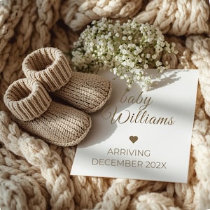 May include: Beige knitted baby booties and a card reading "baby Williams", a heart symbol, and "ARRIVING DECEMBER 202X". The items rest on a cream knitted blanket, with white flowers. The booties are a soft, neutral colour.