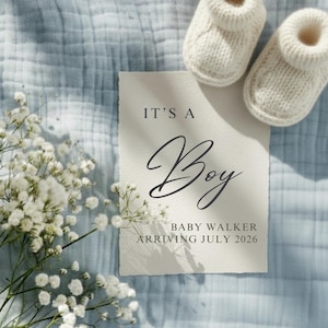 May include: A baby announcement card with the text "IT'S A Boy" in elegant script, along with the baby's last name and expected arrival date. The card is accompanied by a pair of knitted baby booties and white flowers, all on a blue fabric.