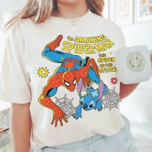 May include: Cream-colored t-shirt featuring a graphic of Spider-Man and Stitch. The text "the AMAZING SPIDER-MAN" is above the characters, with "THE SPIDER OR THE STITCH" below. A white mug with a smiley face is held in the image.