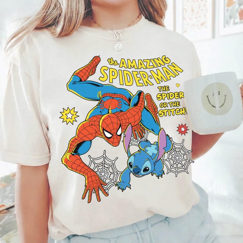 May include: Cream-colored t-shirt featuring a graphic of Spider-Man and Stitch. The text "the AMAZING SPIDER-MAN" is above the characters, with "THE SPIDER OR THE STITCH" below. A white mug with a smiley face is held in the image.