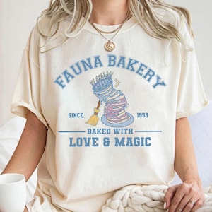 May include: Cream-colored t-shirt with a light blue graphic that reads "Fauna Bakery" above a whimsical cake design. The design includes a broom and the text "Baked with Love & Magic" and "Since 1959".