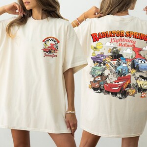 Pode incluir: Camiseta creme com um gr&aacute;fico colorido do filme Carros. A frente tem um pequeno design com um carro de corrida e o nome "Jennifer". A parte de tr&aacute;s exibe "Radiator Springs Lightning McQueen" com v&aacute;rios personagens de carros.