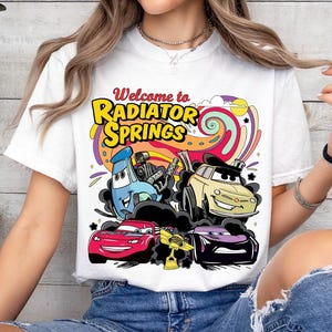 Może przedstawiać: Biała koszulka z kolorową grafiką przedstawiającą kresk&oacute;wkowe samochody i napisem "Welcome to Radiator Springs". Projekt zawiera czerwony samoch&oacute;d, ż&oacute;łty samoch&oacute;d, fioletowy samoch&oacute;d i niebieski samoch&oacute;d.