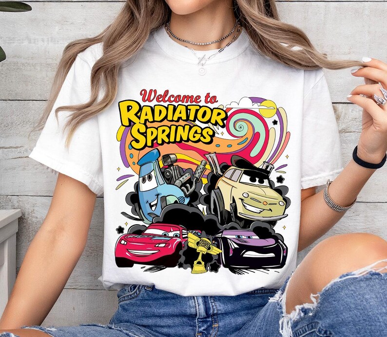Może przedstawiać: Biała koszulka z kolorową grafiką przedstawiającą kresk&oacute;wkowe samochody i napisem "Welcome to Radiator Springs". Projekt zawiera czerwony samoch&oacute;d, ż&oacute;łty samoch&oacute;d, fioletowy samoch&oacute;d i niebieski samoch&oacute;d.