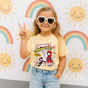 Puede incluir: Una ni&ntilde;a con gafas de sol en forma de coraz&oacute;n y una camiseta amarilla con el texto "Spidey and His Amazing Friends" y gr&aacute;ficos de dibujos animados de Spider-Man y otros personajes. Est&aacute; haciendo el signo de la paz.