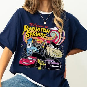 Może przedstawiać: Granatowa koszulka z kolorową grafiką postaci z filmu Auta. Projekt zawiera napis "Welcome to Radiator Springs" w kolorze ż&oacute;łtym i czerwonym, ze zdjęciami samochod&oacute;w i psychodelicznym tłem.