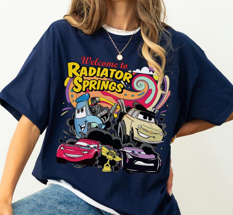 Może przedstawiać: Granatowa koszulka z kolorową grafiką postaci z filmu Auta. Projekt zawiera napis "Welcome to Radiator Springs" w kolorze ż&oacute;łtym i czerwonym, ze zdjęciami samochod&oacute;w i psychodelicznym tłem.