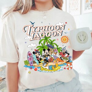 May include: Cream-colored t-shirt featuring a colorful graphic of Disney characters at Typhoon Lagoon. The design includes Mickey Mouse, Minnie Mouse, Donald Duck, Daisy Duck, and Goofy on a tropical island with palm trees, a sun, and the words "Typhoon Lagoon". A white mug with a smiley face is also visible.