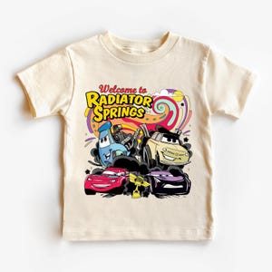 Może przedstawiać: Kremowy t-shirt z kolorową grafiką samochod&oacute;w z kresk&oacute;wek z napisem "Welcome to Radiator Springs". Projekt zawiera czerwony samoch&oacute;d, beżowy samoch&oacute;d, fioletowy samoch&oacute;d i niebieski samoch&oacute;d.