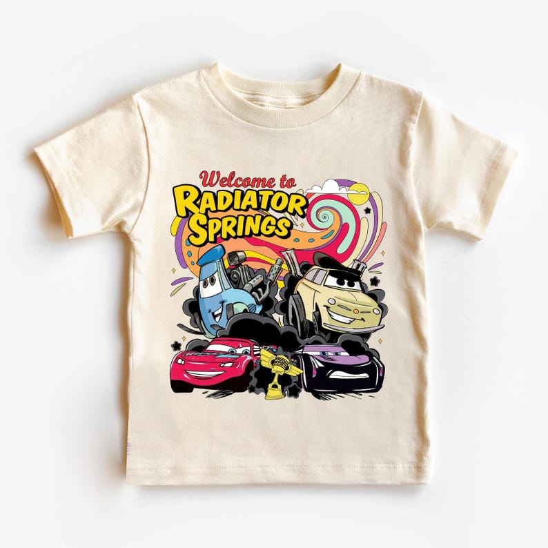 Może przedstawiać: Kremowy t-shirt z kolorową grafiką samochod&oacute;w z kresk&oacute;wek z napisem "Welcome to Radiator Springs". Projekt zawiera czerwony samoch&oacute;d, beżowy samoch&oacute;d, fioletowy samoch&oacute;d i niebieski samoch&oacute;d.