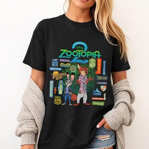 Op de afbeelding: Zwart T-shirt met een ontwerp uit de Disney-film "Zootopia". Het ontwerp bevat de titel van de film, personages en stadsborden. Het T-shirt heeft een ronde hals en korte mouwen.