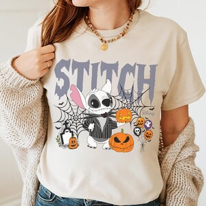 Pode incluir: Camiseta bege com um design de Halloween com Stitch vestido como Jack Skellington, ab&oacute;boras, teias de aranha e a palavra "STITCH" em roxo. O logotipo da Disney &eacute; vis&iacute;vel. A camiseta &eacute; de gola redonda e manga curta.