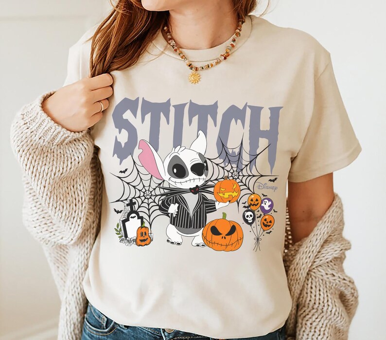 Pode incluir: Camiseta bege com um design de Halloween com Stitch vestido como Jack Skellington, ab&oacute;boras, teias de aranha e a palavra "STITCH" em roxo. O logotipo da Disney &eacute; vis&iacute;vel. A camiseta &eacute; de gola redonda e manga curta.