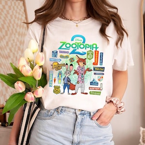 Op de afbeelding: Wit T-shirt met een kleurrijke afbeelding uit de Disney-film Zootopia. Het ontwerp bevat de titel van de film, de personages Judy Hopps en Nick Wilde en diverse thematische borden. Het T-shirt wordt door een persoon gedragen.