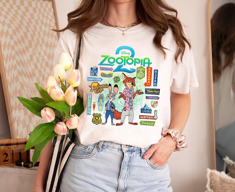 Op de afbeelding: Wit T-shirt met een kleurrijke afbeelding uit de Disney-film Zootopia. Het ontwerp bevat de titel van de film, de personages Judy Hopps en Nick Wilde en diverse thematische borden. Het T-shirt wordt door een persoon gedragen.