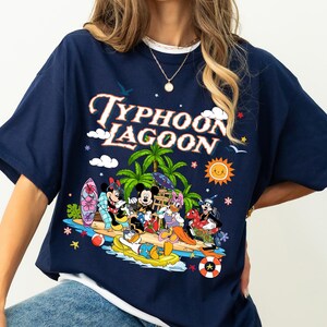 May include: Navy blue t-shirt featuring the words "Typhoon Lagoon" above a colorful graphic of Disney characters at a tropical beach. The graphic includes Mickey Mouse, Minnie Mouse, Donald Duck, and Goofy.