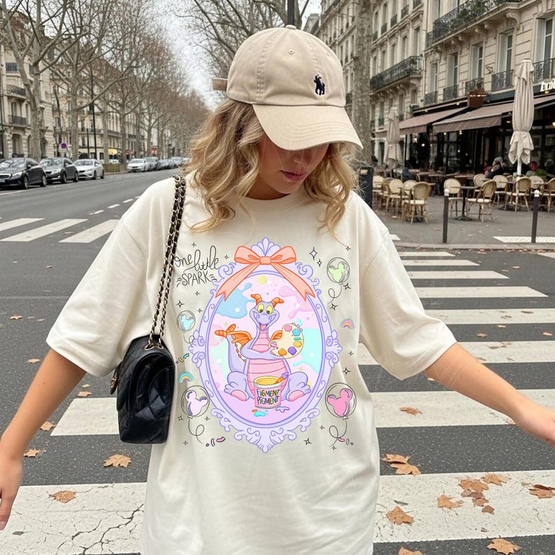 May include: Cream-colored t-shirt featuring a whimsical illustration of a purple dragon holding a paintbrush and palette. The shirt has the text "one little spark" and "Figment Promen" in a decorative frame. A beige baseball cap completes the look.
