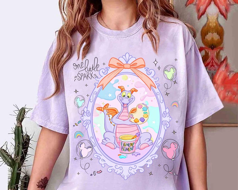 May include: A light purple t-shirt featuring a cartoon dragon inside an ornate frame. The dragon holds a palette and paint, with the words "one little SPARK" above. The design includes a bow, stars, and Mickey Mouse-shaped balloons.